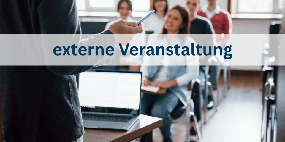 Im Hintergrund ist verschwommen eine Klasse mit erwachsenen menschen zu sehen. Vorne steht ein Dozent mit einem aufgeklappten Laptop auf dem Tisch.
