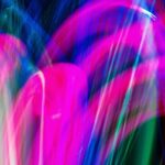 Das Bild zeigt eine bunte abstrakte Lichtmalerei. Es gibt keine erkennbaren Figuren oder Gegenstände. Stattdessen sieht man geschwungene Linien und Bögen in leuchtenden Farben: Pink / Magenta dominiert das Bild mit kräftigen Bögen. Blau ist stark im Hintergrund und an den Rändern zu sehen. Grün erscheint als einzelne vertikale und geschwungene Linien dazwischen. Stellenweise mischen sich die Farben ineinander, sodass fließende Übergänge entstehen. Das Bild wirkt lebendig, dynamisch und erinnert an eine Lichtshow, Neonfarben oder lange Belichtungszeiten bei bewegtem Licht.