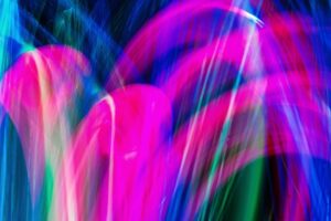 Das Bild zeigt eine bunte abstrakte Lichtmalerei. Es gibt keine erkennbaren Figuren oder Gegenstände. Stattdessen sieht man geschwungene Linien und Bögen in leuchtenden Farben: Pink / Magenta dominiert das Bild mit kräftigen Bögen. Blau ist stark im Hintergrund und an den Rändern zu sehen. Grün erscheint als einzelne vertikale und geschwungene Linien dazwischen. Stellenweise mischen sich die Farben ineinander, sodass fließende Übergänge entstehen. Das Bild wirkt lebendig, dynamisch und erinnert an eine Lichtshow, Neonfarben oder lange Belichtungszeiten bei bewegtem Licht.